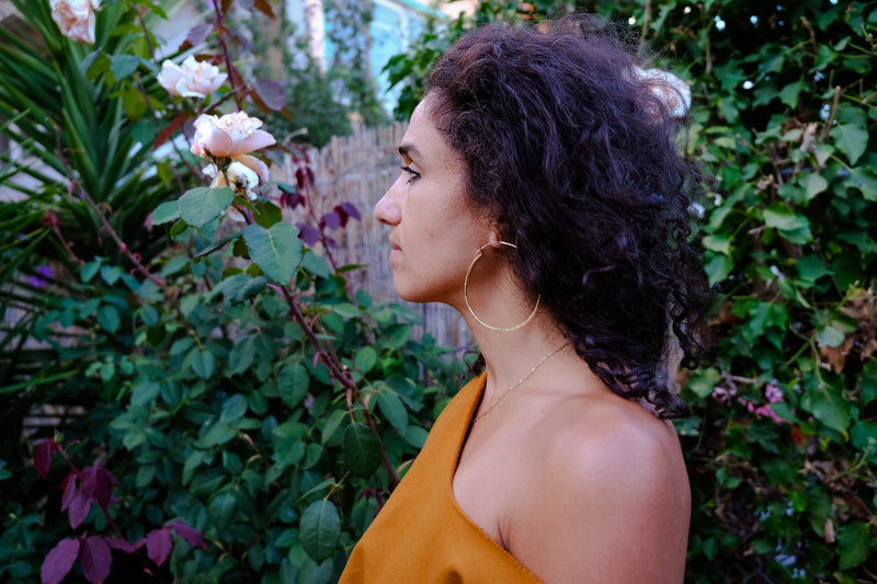 naima wearing 3 inch thin brass hoops in a garden with green plants and several peach colored roses
